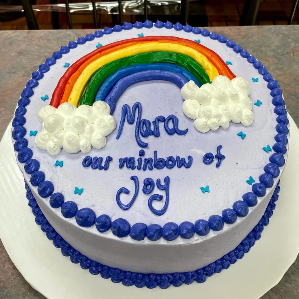 A round cake with light purple frosting, a rainbow, clouds, and "Mara our rainbow of Joy" written in blue.