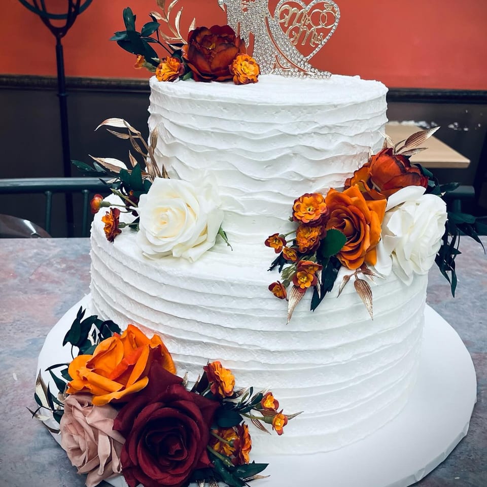Two-tier white wedding cake with textured frosting, adorned with orange, red, and white flowers.