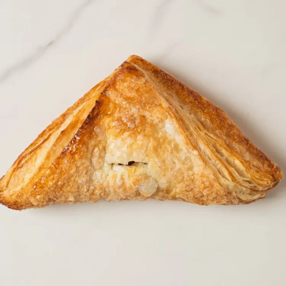 A golden-brown, triangular Elmwood Pastry Shop apple turnover with flaky crust on a white surface.
